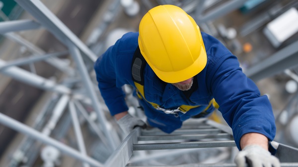 A high-angle view looking down at a construction worker in a blue uniform and yellow hard hat climbing a metal ladder on a high-altitude structure in New York City. The background shows a blurred view of the ground far below. (1845662835)