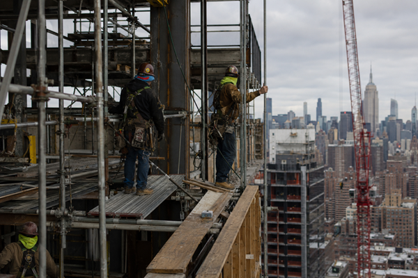 Construction workers in hard hats and safety harnesses work on open scaffolding high above the New York City skyline with the Empire State Building visible in the background, depicting the fall hazard and OSHA safety violation risks facing New York construction workers.