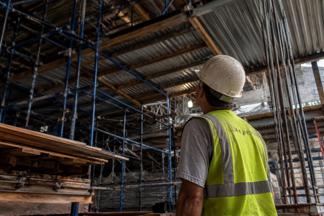 A construction worker wearing a white hard hat and a high-visibility safety vest looks up at a massive, complex network of scaffolding and exposed overhead building structures.